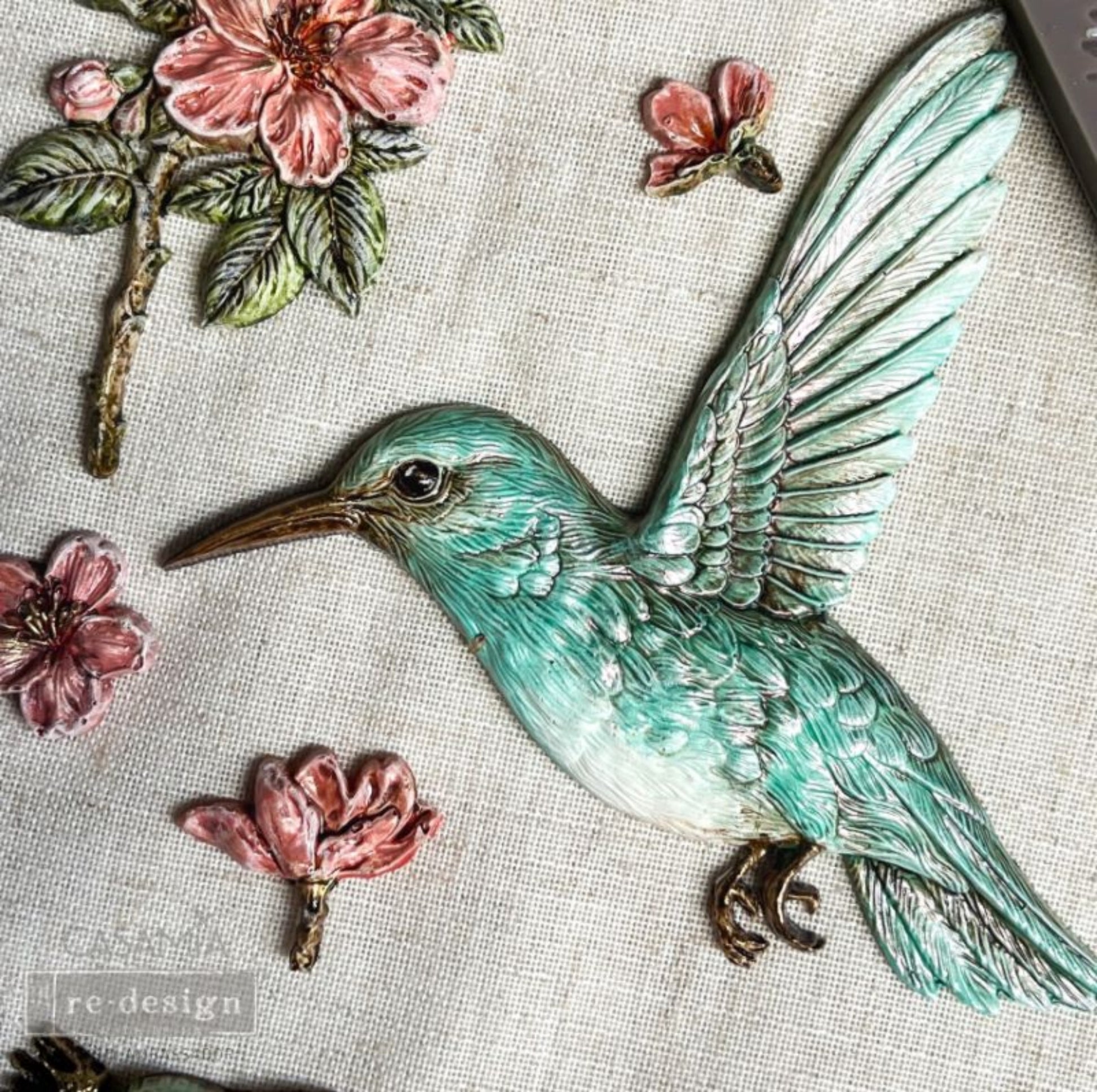 Close-up of Bloom & Flutter silicone mould with detailed hummingbird feathers, delicate flowers, leafy branches, and small crown accents.