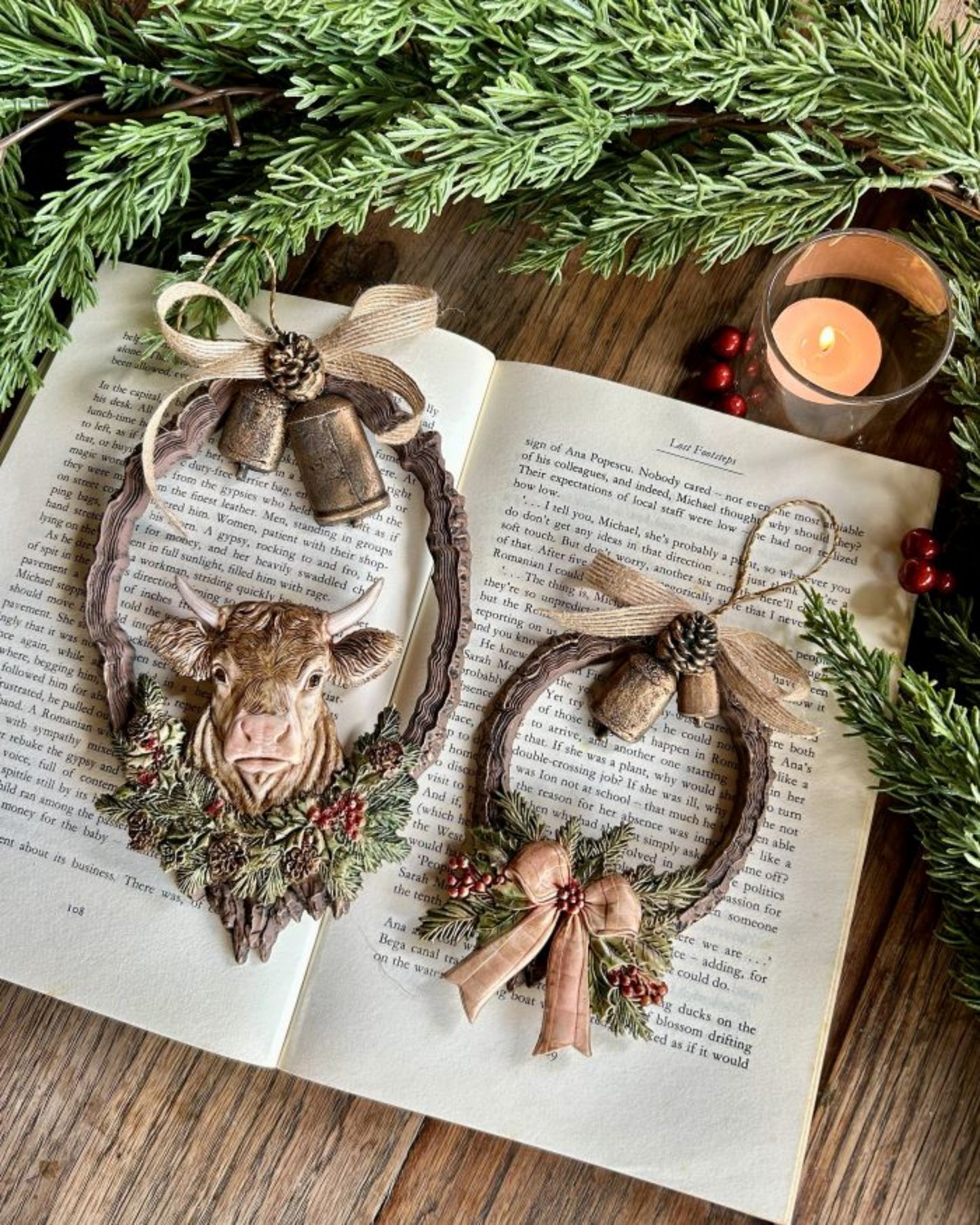 Two rustic holiday wreath ornaments featuring cow, bell, bow, evergreen, and pinecone castings displayed on an open book.