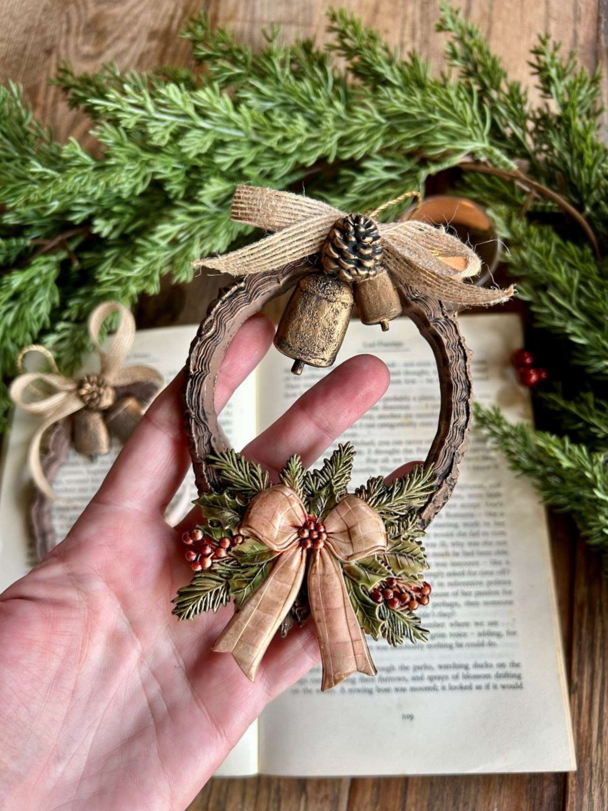 Hand holding a finished woodland wreath ornament made with tree-ring, bell, bow, evergreen, berry, and pinecone castings.