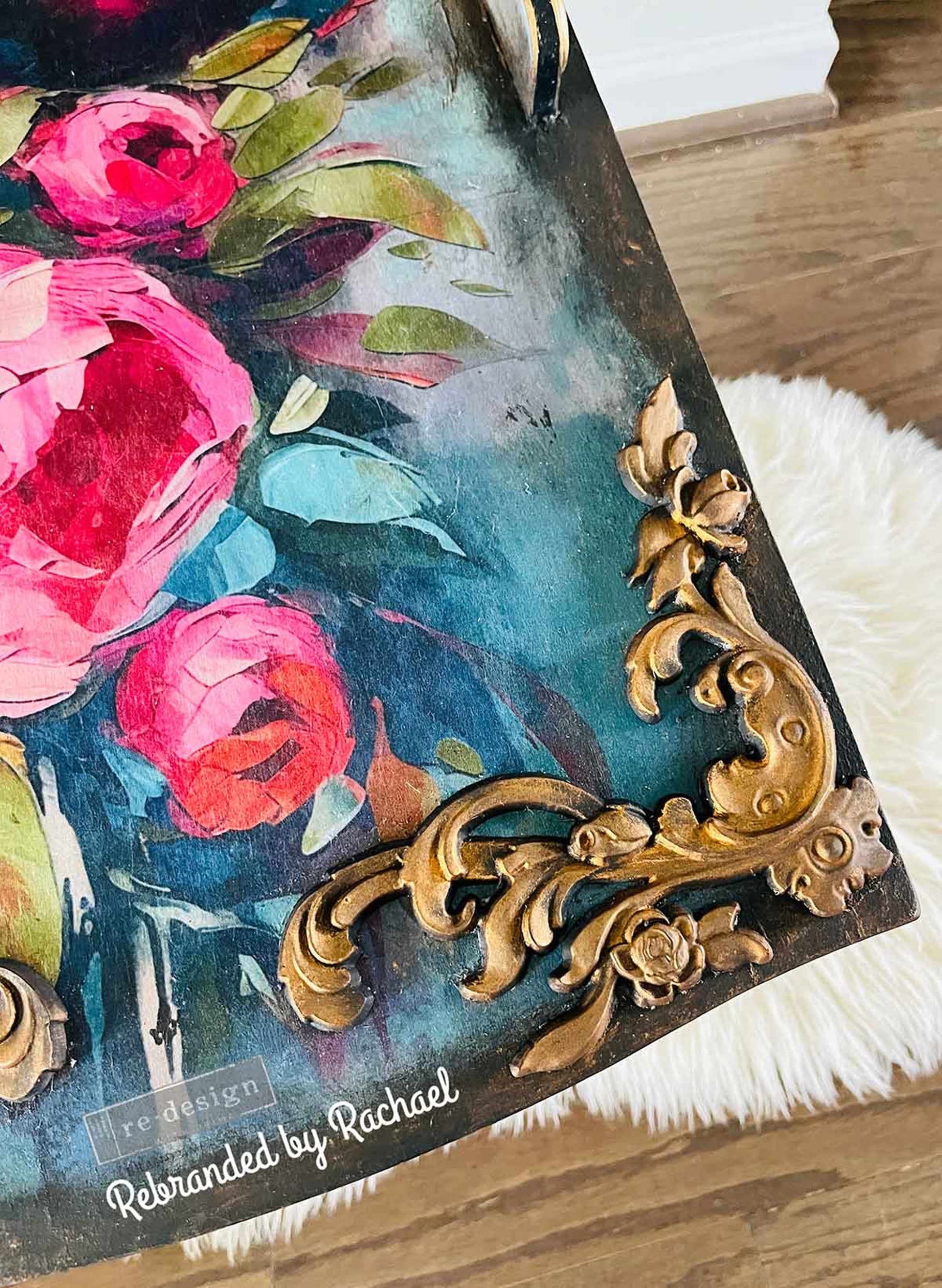 Intricate Grace Décor Poly® appliqué finished in antique gold and applied to the corner of a dark floral furniture piece with vibrant pink roses and teal accents.