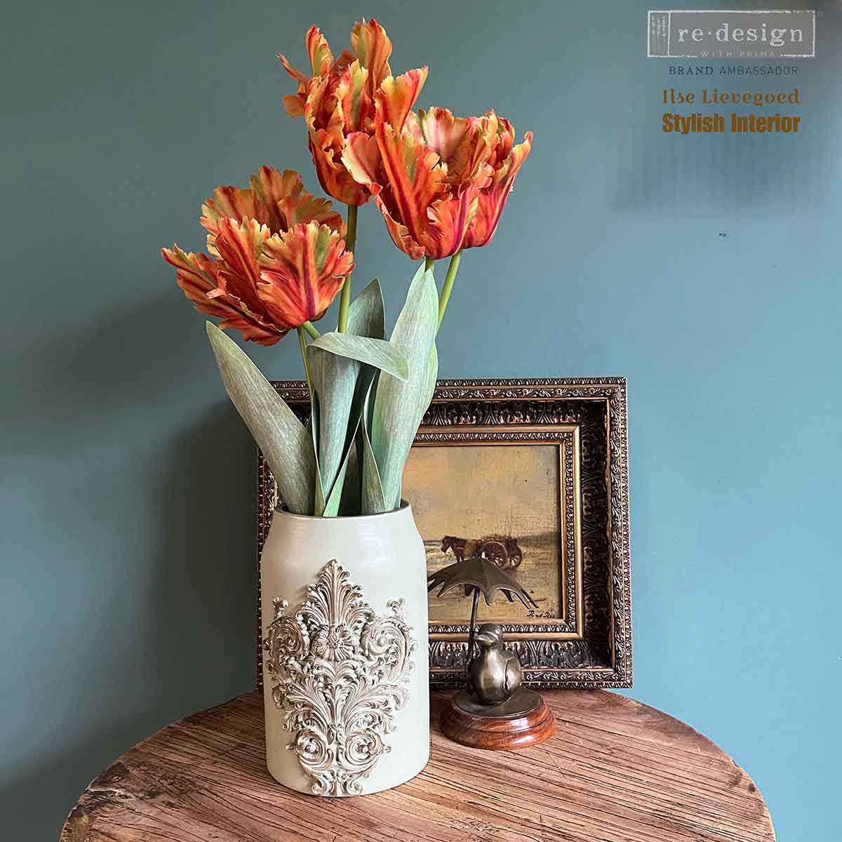 Salon De La Belle Epoque mould casting used on a cream vase with raised ornamental detail, styled with orange tulips and vintage framed art.